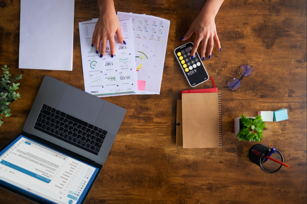 Overhead view of a businesswoman analyzing financial documents on a wooden desk with digital tools.